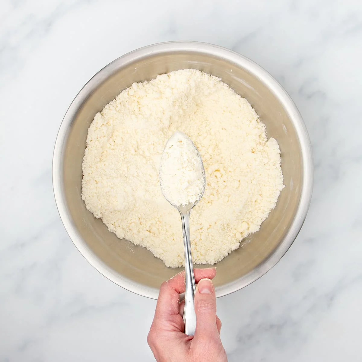 Hand holding metal mixing spoon with sandy pale tan mixture of butter mixed with flour, and more of the mixture in the same mixing bowl.