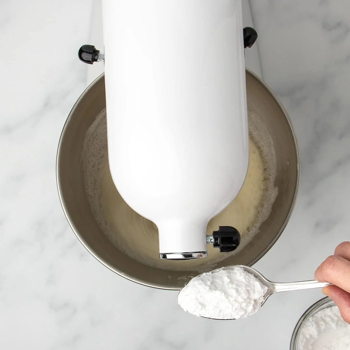 hand holding metal mixing spoon with white flour mixture next to white stand mixer beating egg whites