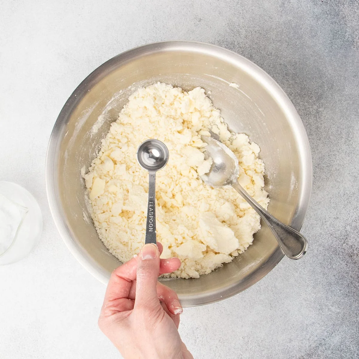 Hand holding metal teaspoon with water about to be poured into mixing bowl with slightly dry somewhat clumpy light tan pastry mixture.