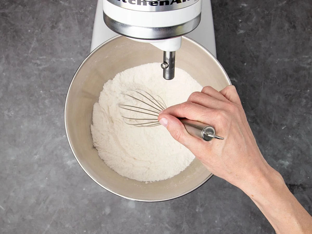 hand holding metal wire whisk, whisking white flour mixture in bowl of stand mixer