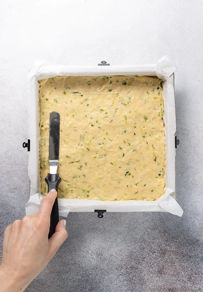 Hand holding offset spatula spreading zucchini cake batter in square pan lined with white paper