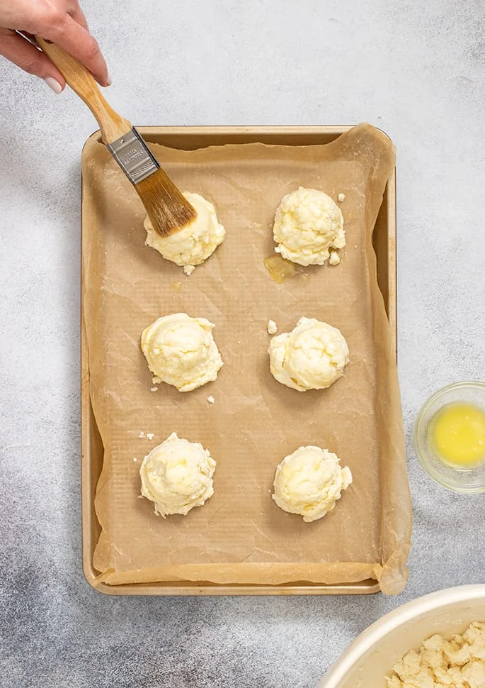Hand holding pastry brush brushing melted butter on one of 6 raw biscuits on tray lined with brown paper.