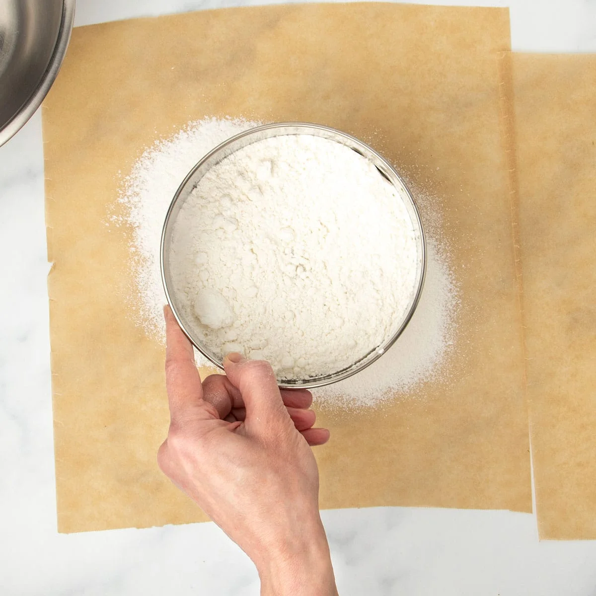 hand holding round metal mesh sieve with white flour over brown sheet of parchment paper