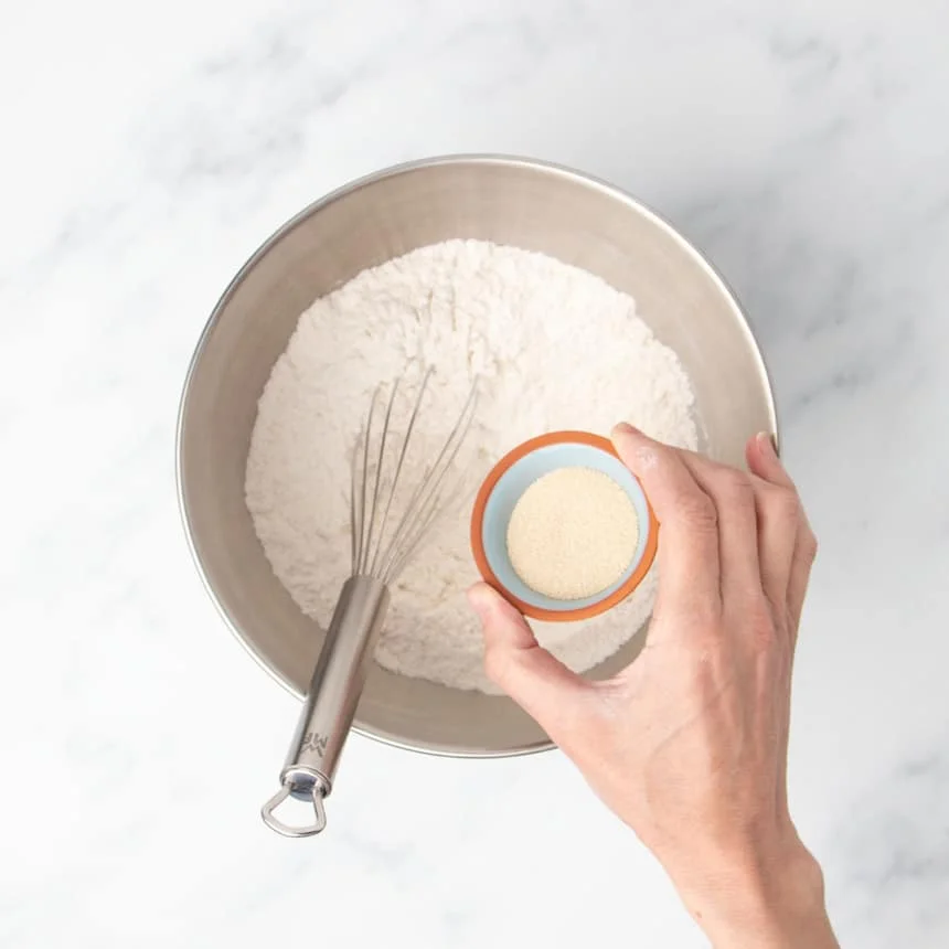 hand holding small blue bowl with light brown yeast granules over a large metal round mixing bowl with flour and a wire whisk