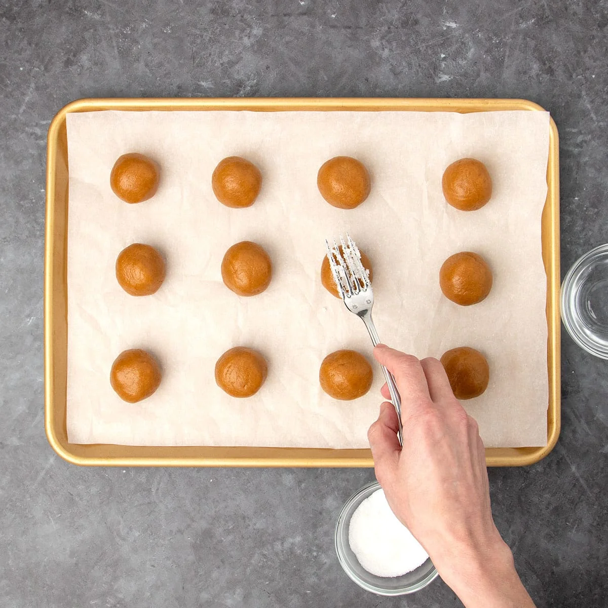 hand holding small metal fork with white sugar stuck to the tines, positioned over ball of cookie dough about to make a mark on the dough