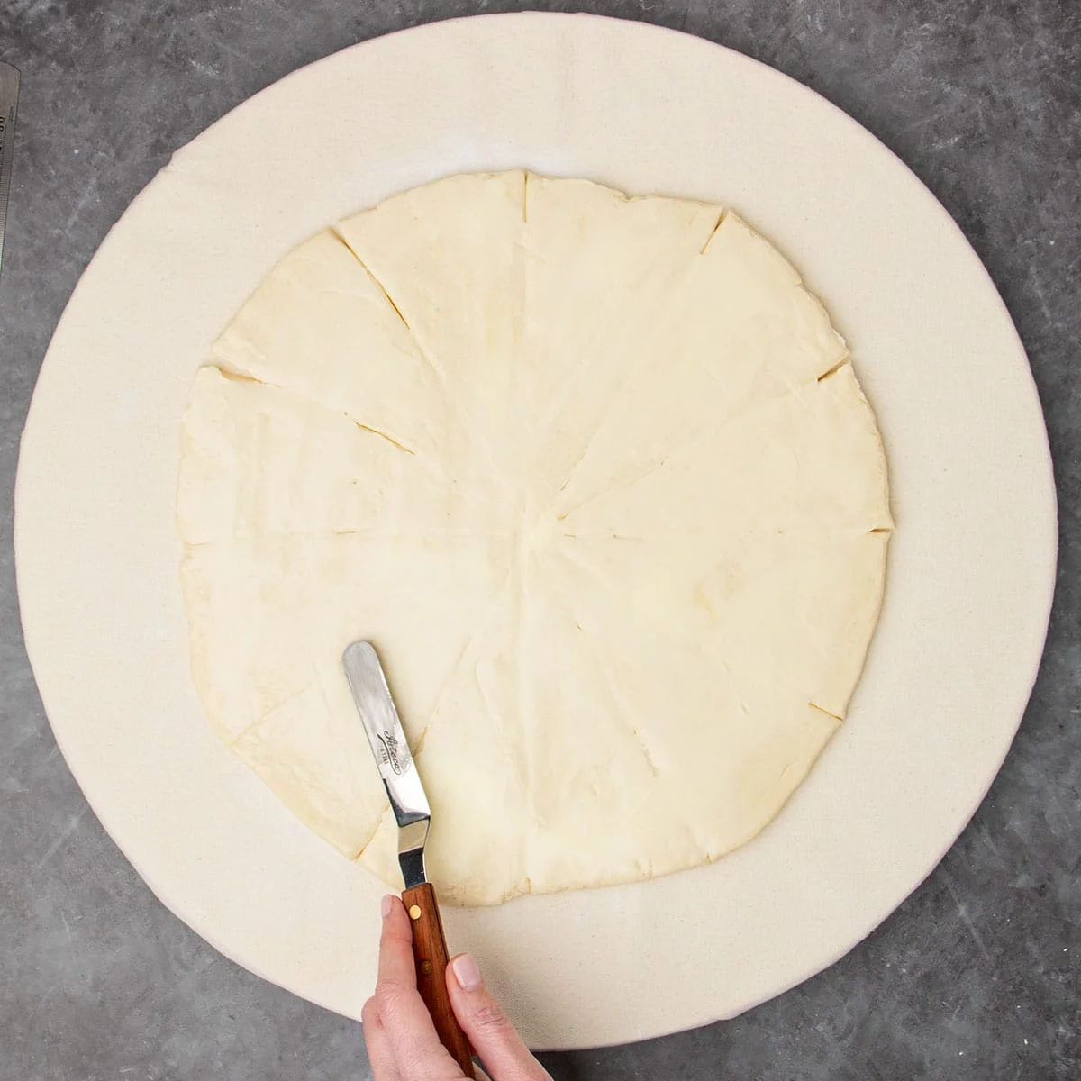 hand holding small offset metal spatula with brown handle spreading soft butter on same sliced dough round