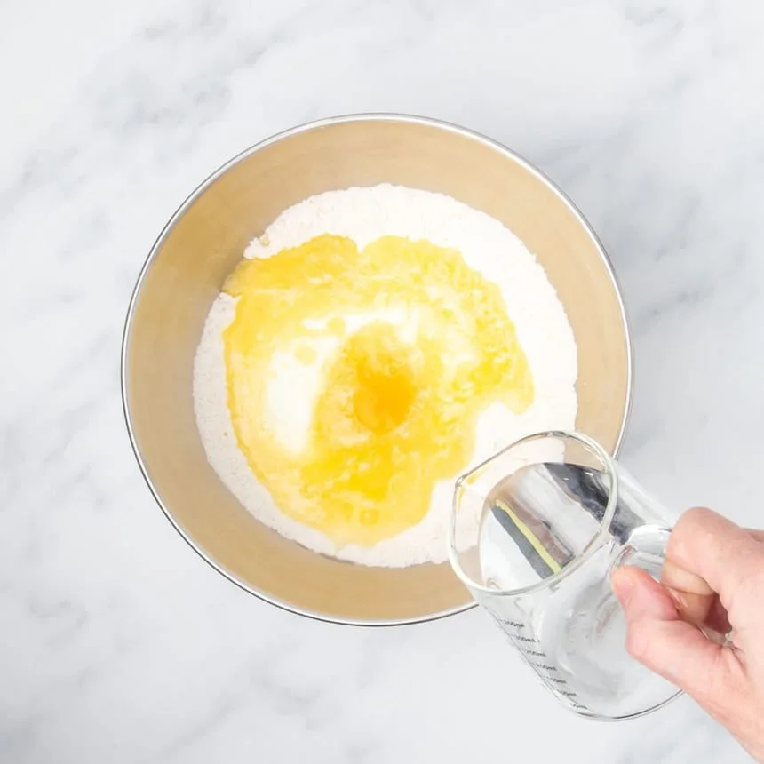 hand holding small pitcher pouring water into same mixing bowl with yellow egg and melted butter and white yogurt on top of flour in bowl