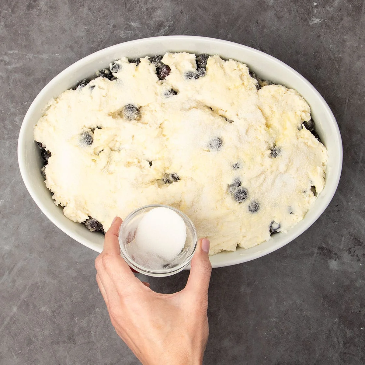 hand holding very small glass dish sprinkling white granulated sugar on top of topping in white dish