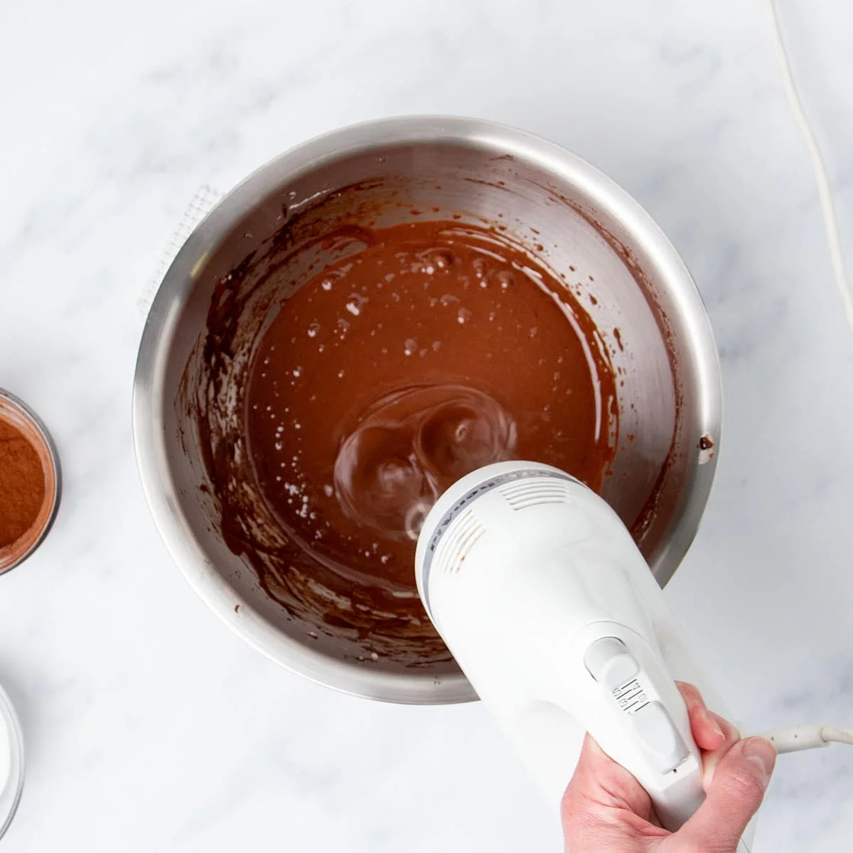 Hand holding white handheld mixer beating egg whites with cocoa powder and sugar.