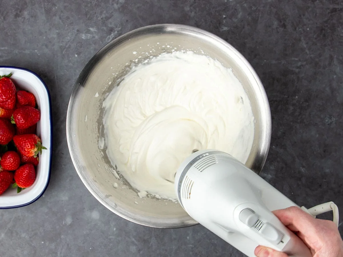 hand holding white handheld mixer being used to whip white cream into light and fluffy whipped cream in round metal mixing bowl with red strawberries in white dish on the side