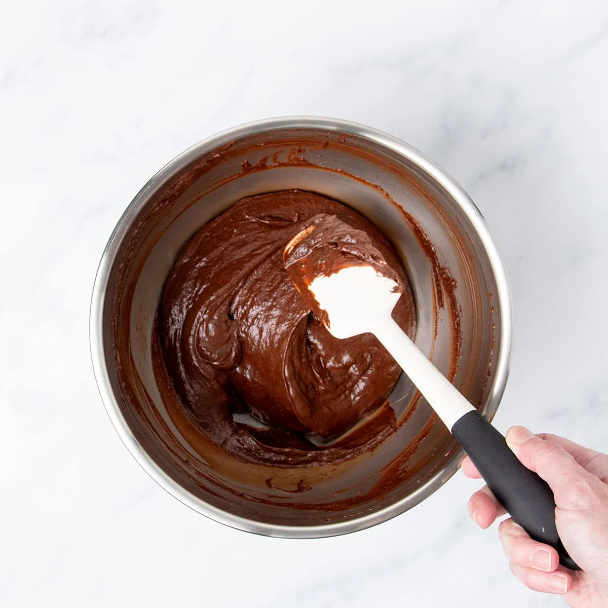 Hand holding white spatula folding in chocolate meringue mixture into whisked ingredients in same bowl.