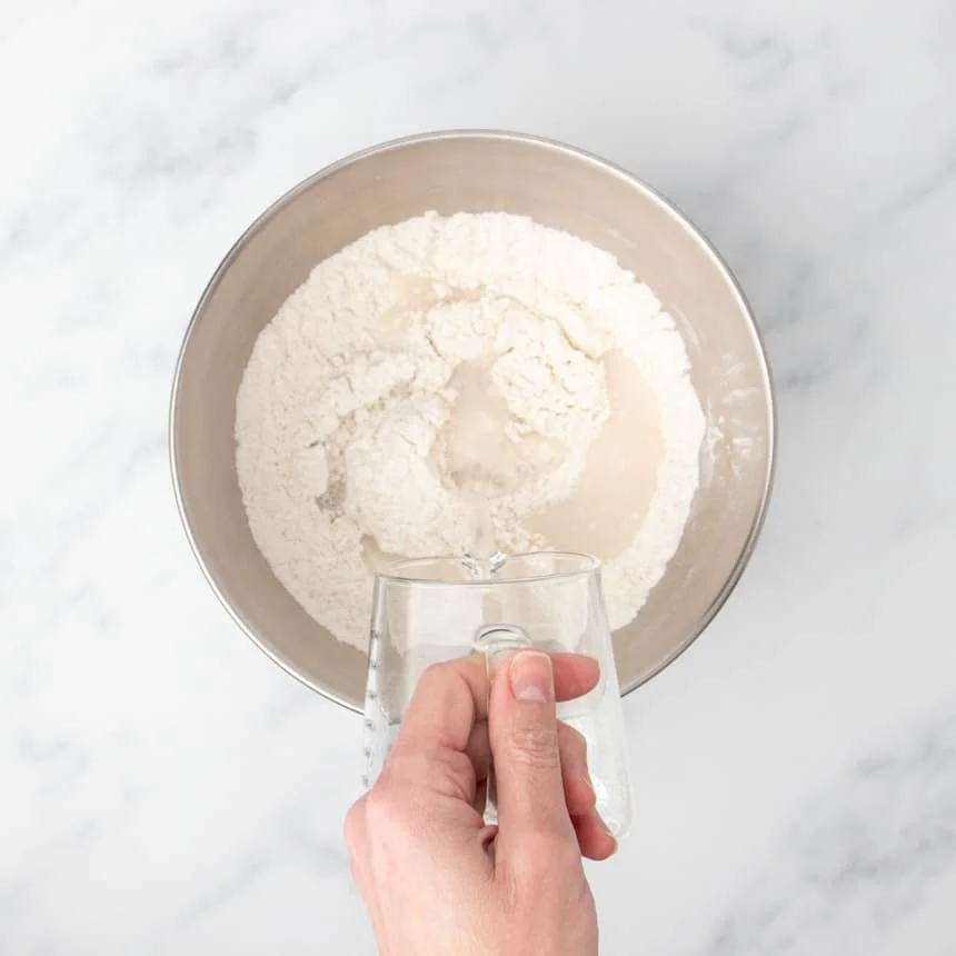 hand pouring water from a small glass pitcher into the same large metal mixing bowl with whisked flour and other dry ingredients
