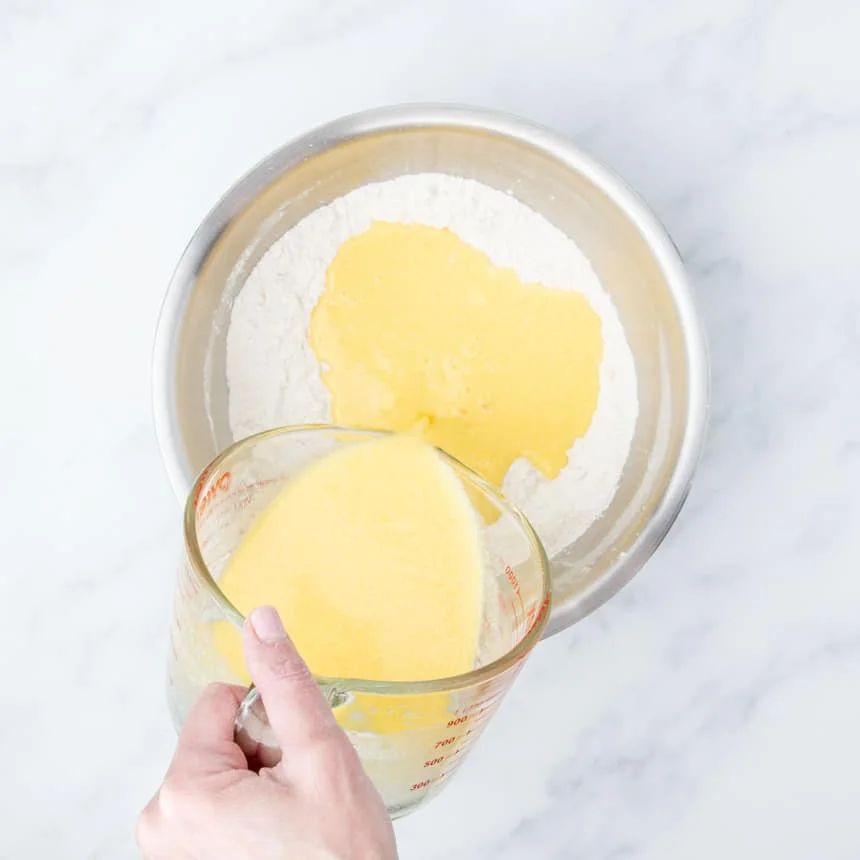 hand pouring yellow liquid from glass measuring cup into large round metal mixing bowl with whisked white flour mixture inside