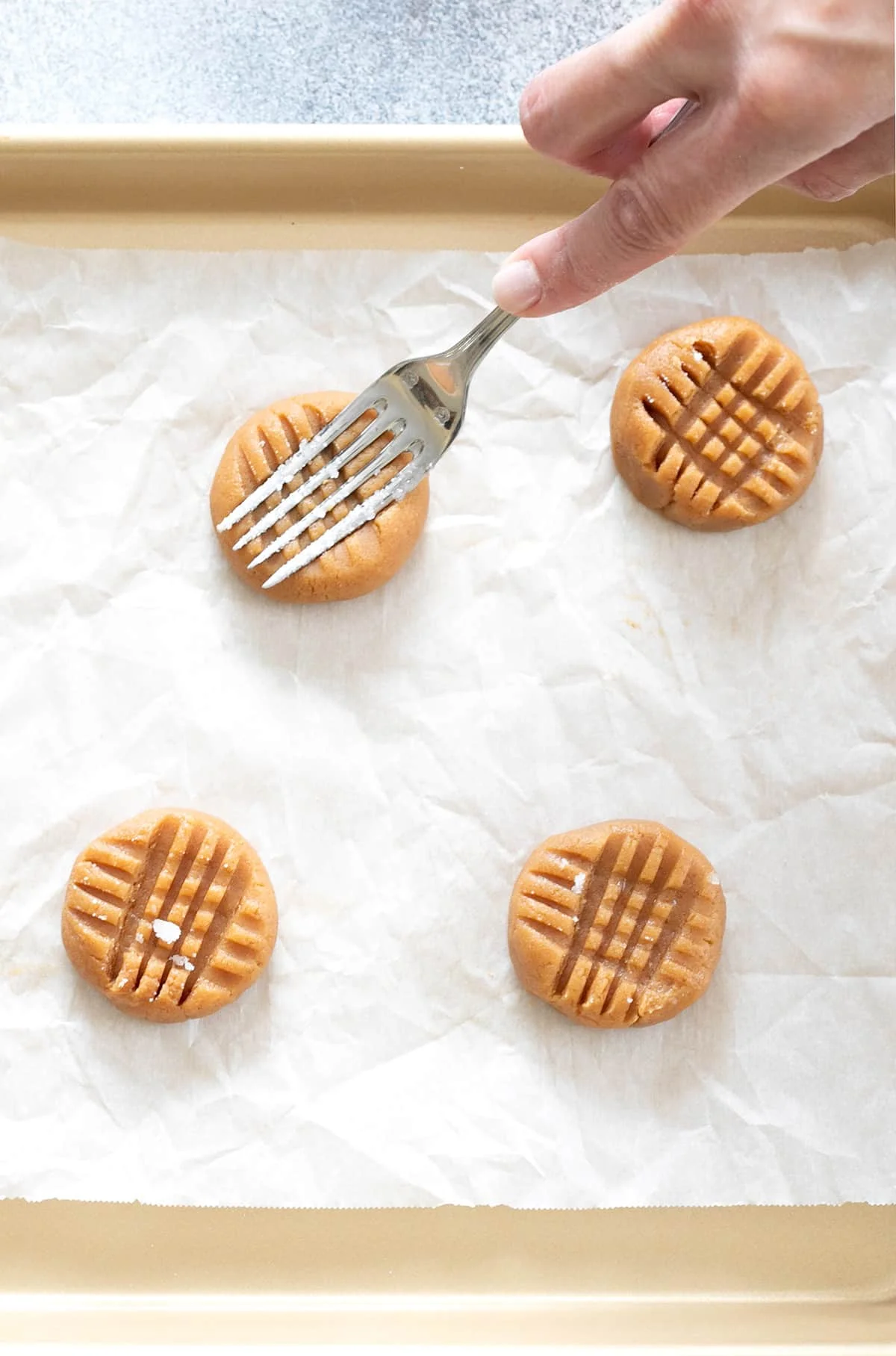 Hand pressing a fork into flourless peanut butter cookie dough on white paper on gold colored baking sheet