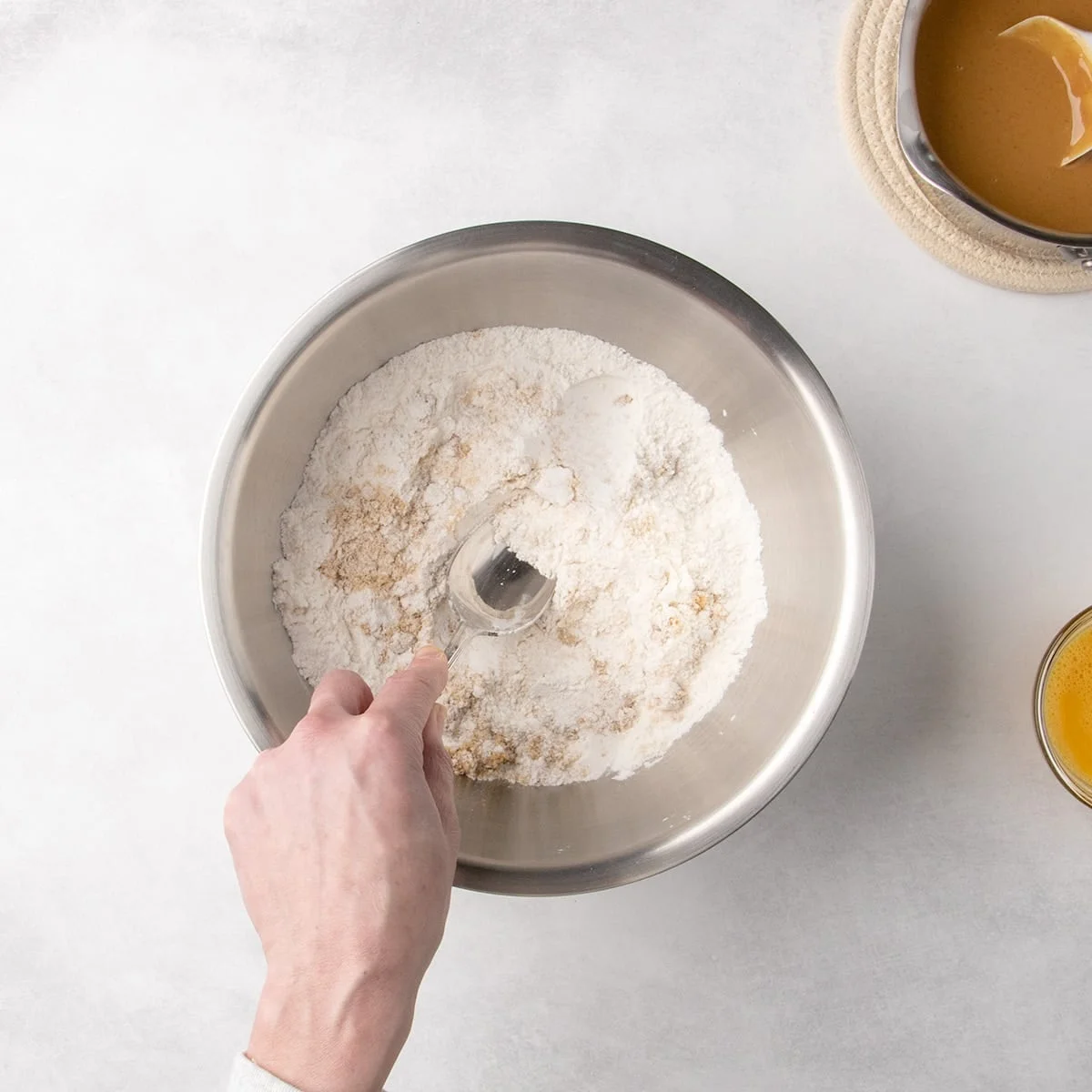 Hand pressing down with back of spoon into a metal mixing bowl containing white dry ingredients and brown sugar.
