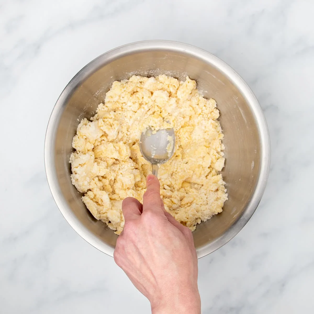 Hand pressing metal mixing spoon into sandy mixture in bowl with beaten egg added, pressing mixture into clumps.