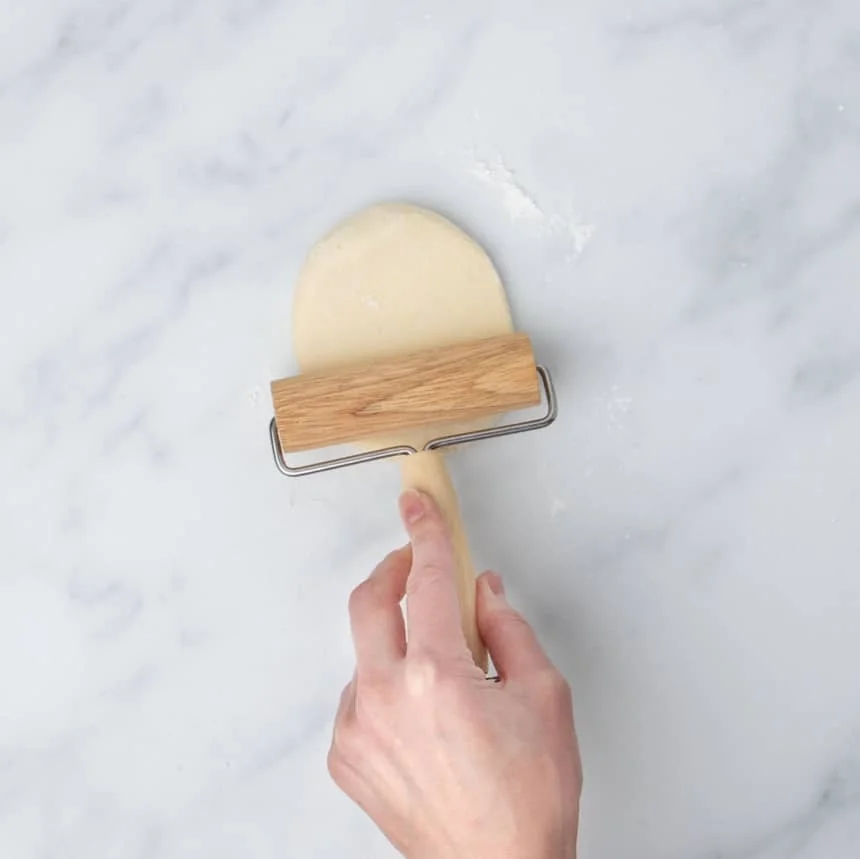 hand pressing small blond wood roller into ball of dough to create an oval shape