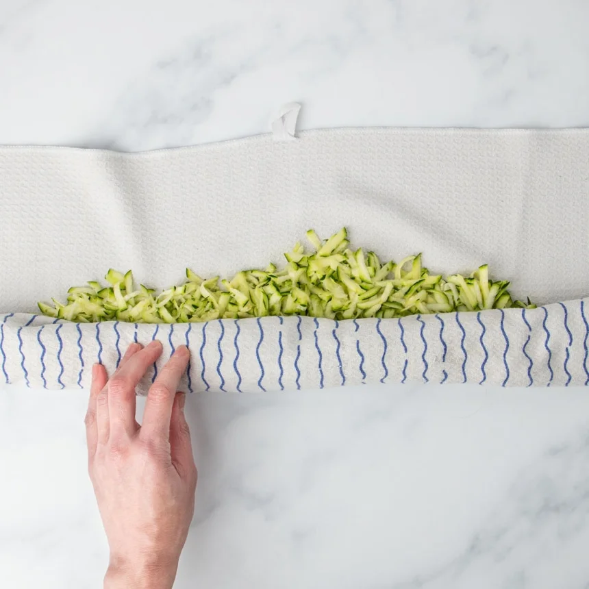 hand rolling white towel with blue stripes over shredded green zucchini