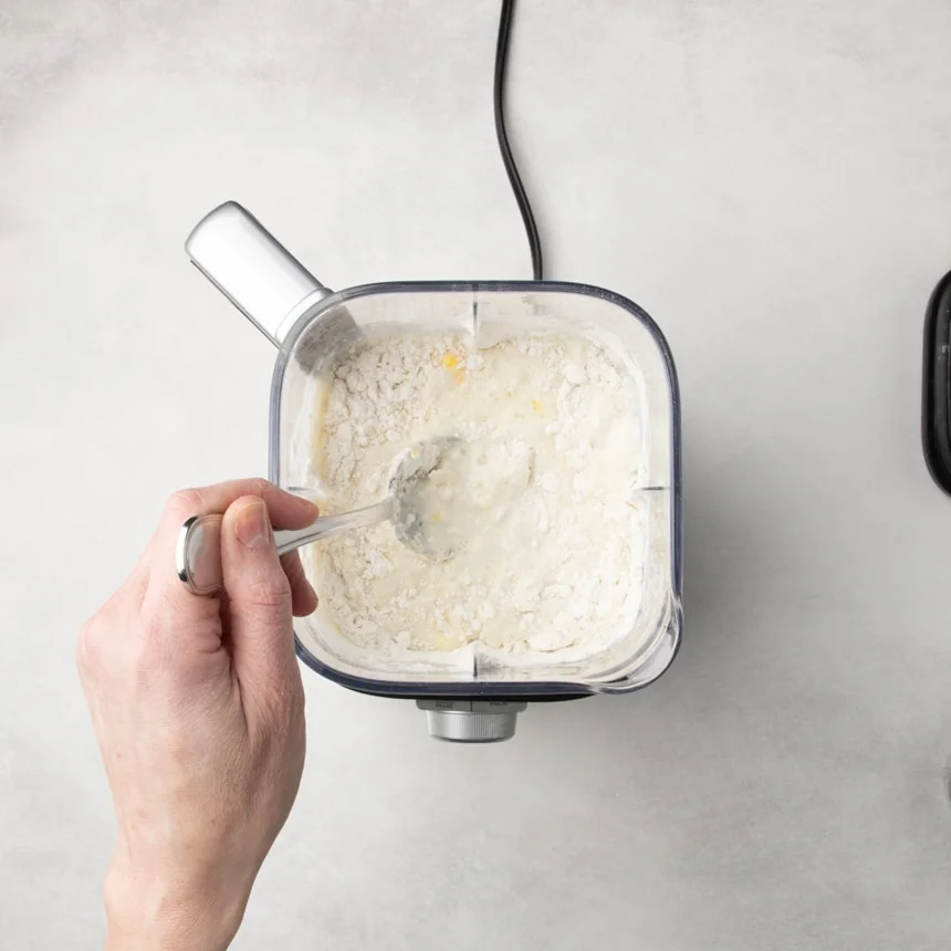 Hand stirring flour and liquid together inside a blender container with a spoon.