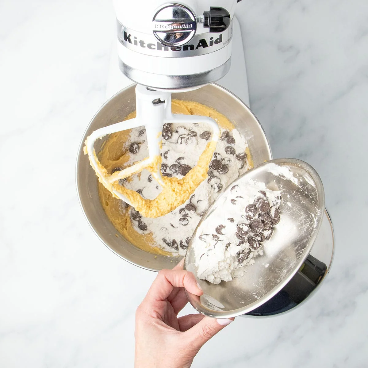 Hand tipping metal bowl with gluten free flour and chocolate chips into soft light brown dough in the mixing bowl with a stand mixer paddle attachment.