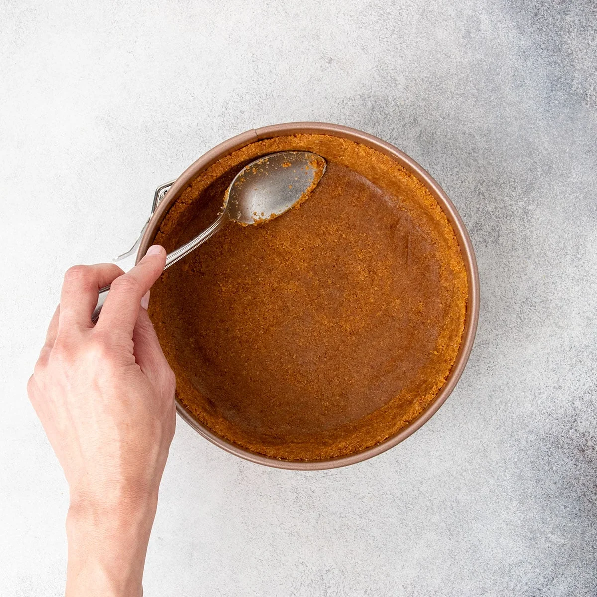 Hand using large metal mixing spoon to pat down brown clumpy crumb mixture into bottom and up sides of pan.