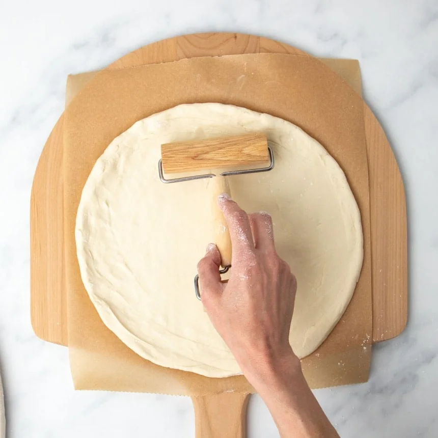 hand using small light blond wood pizza roller to press same dough into a large, smooth round, with dough now on brown paper on a blond wood pizza peel