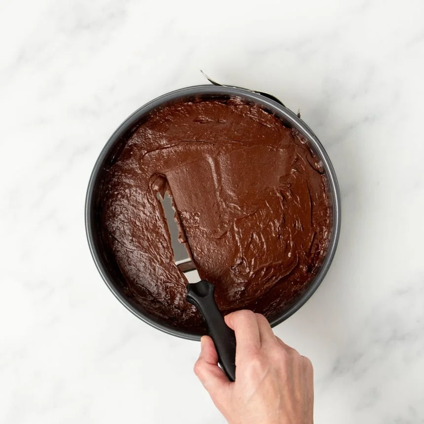 Hand using small metal offset spatula with black handle to spread out raw flourless chocolate cake batter in same round springform pan.