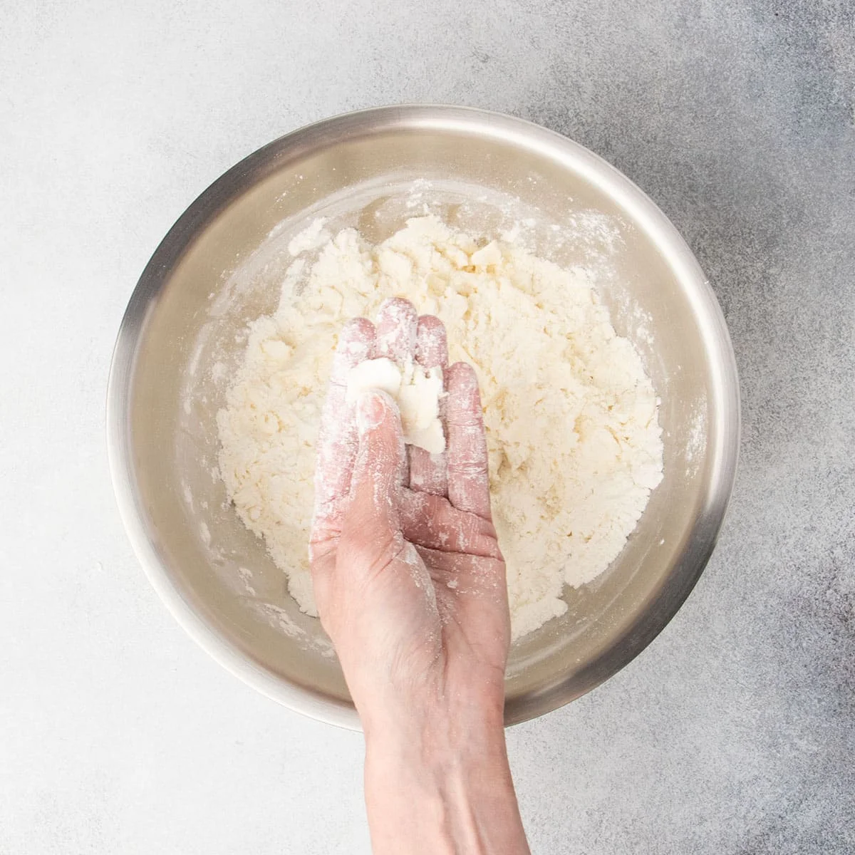 Hand with flour on fingers pressing pieces of butter covered in flour flat between thumb and fingers, held over mixing bowl.