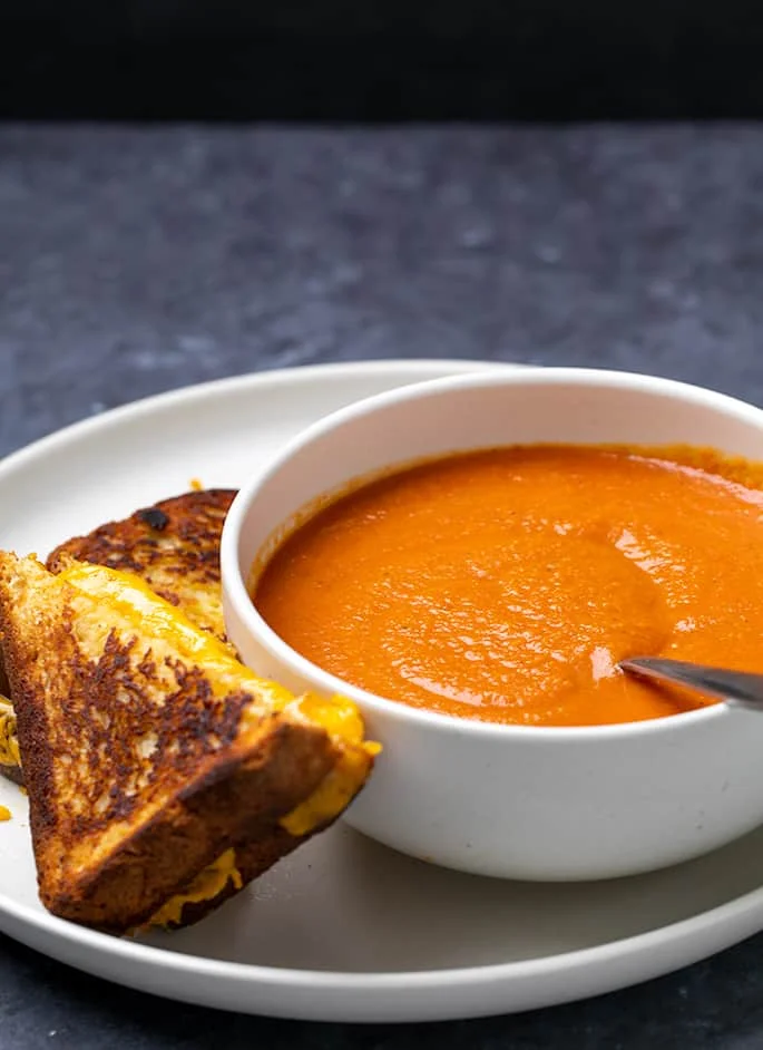 Handle of spoon coming out of bowl of tomato soup with grilled cheese triangles on the side on a plate