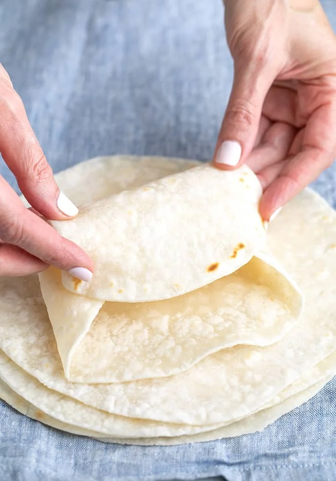 Hands folding top gluten free tortilla on a stack into a burrito shape, on blue cloth