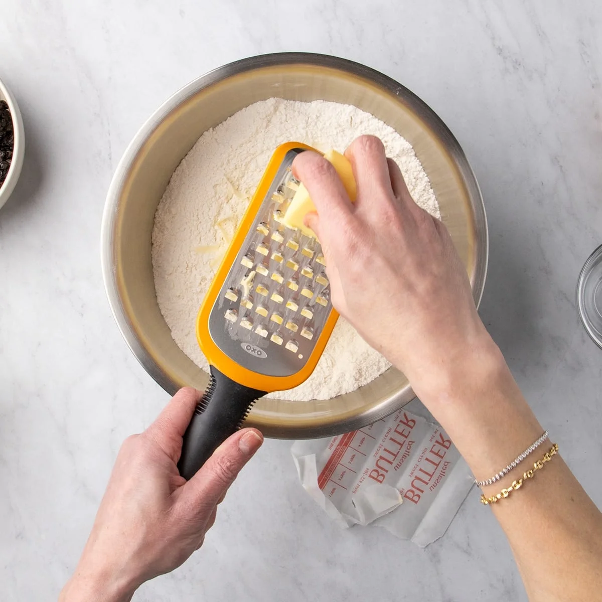 Hands grating a stick of butter on a coarse grater directly into a bowl of white dry ingredients.