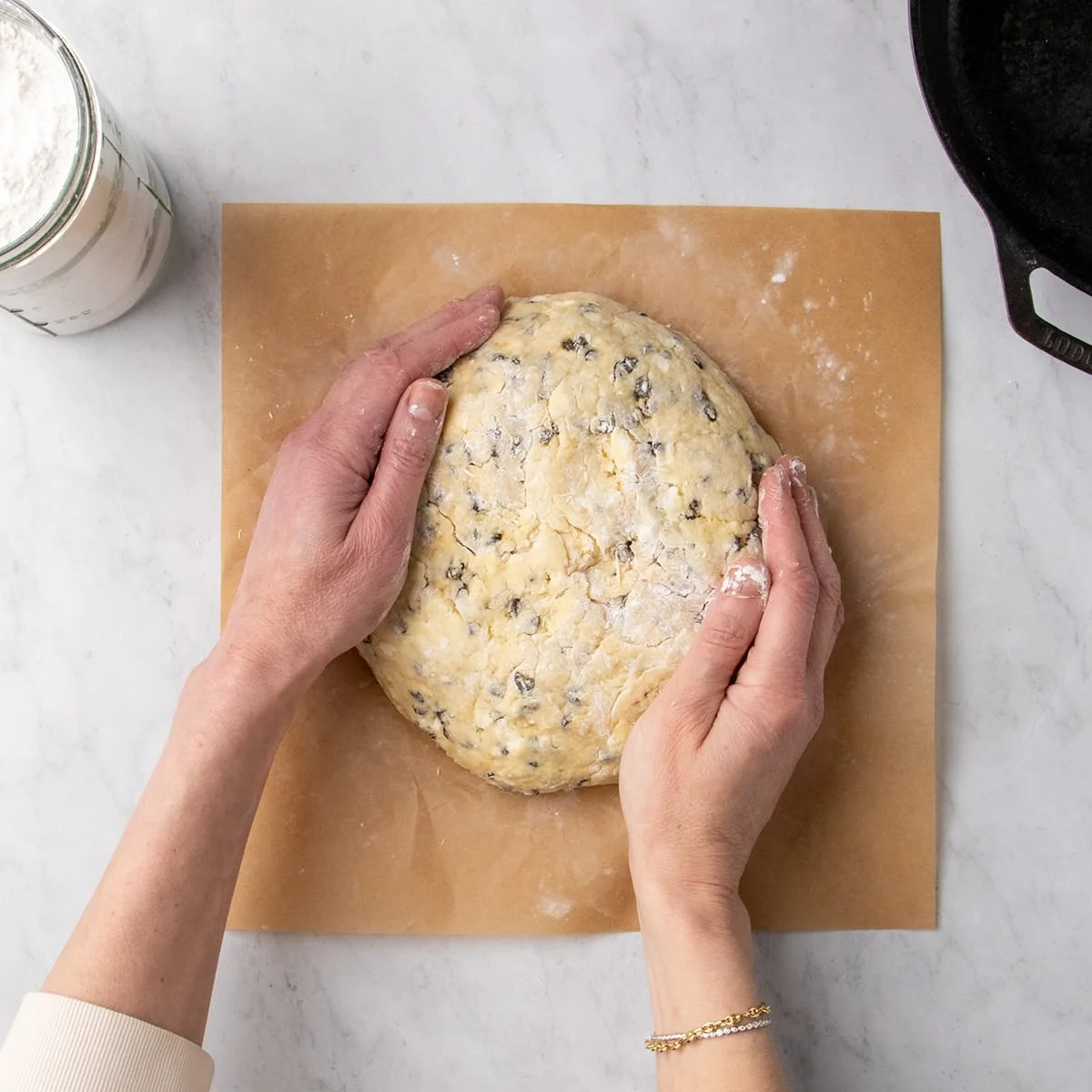 Hands shaping a round of raw soda bread dough with small dark raisins on brown parchment paper on a marble surface.