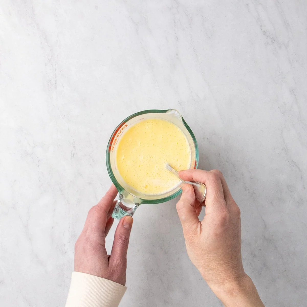 Hands stirring a pale yellow liquid in a glass measuring cup with a metal fork on a marble surface.