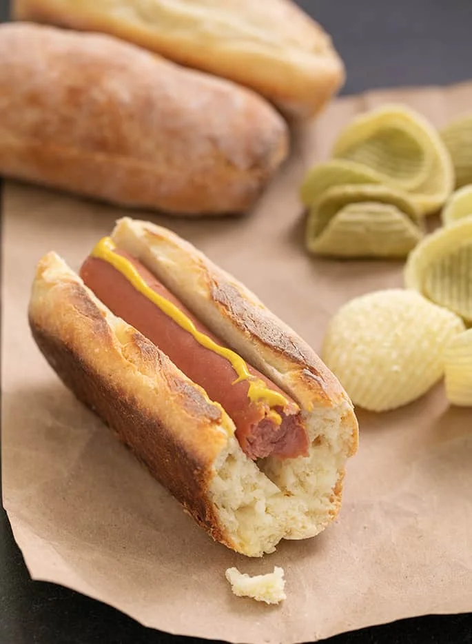 Hot dog with mustard has a bite out, served on brown paper with chips.