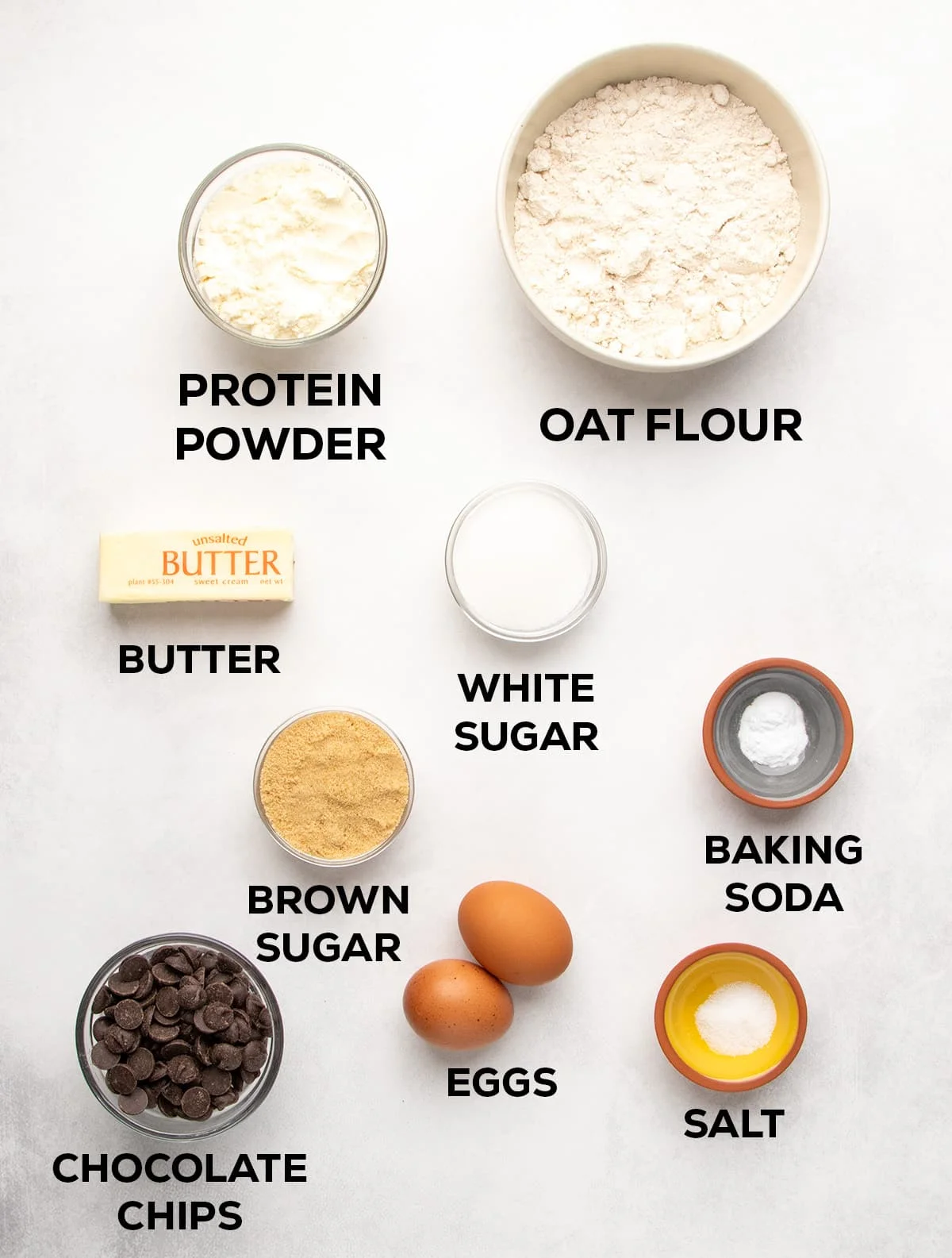 Ingredients arranged on a light surface with text labels: protein powder, oat flour, butter, white sugar, baking soda, brown sugar, eggs, salt, and chocolate chips.