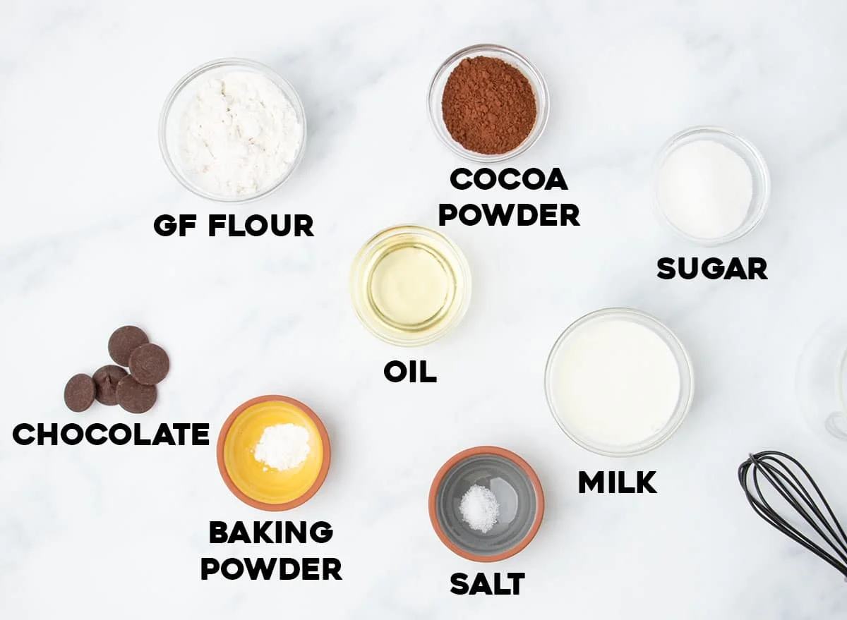 Ingredients for a gluten free mug cake, labeled and arranged on a white surface, including flour, cocoa, chocolate, oil, milk, and more