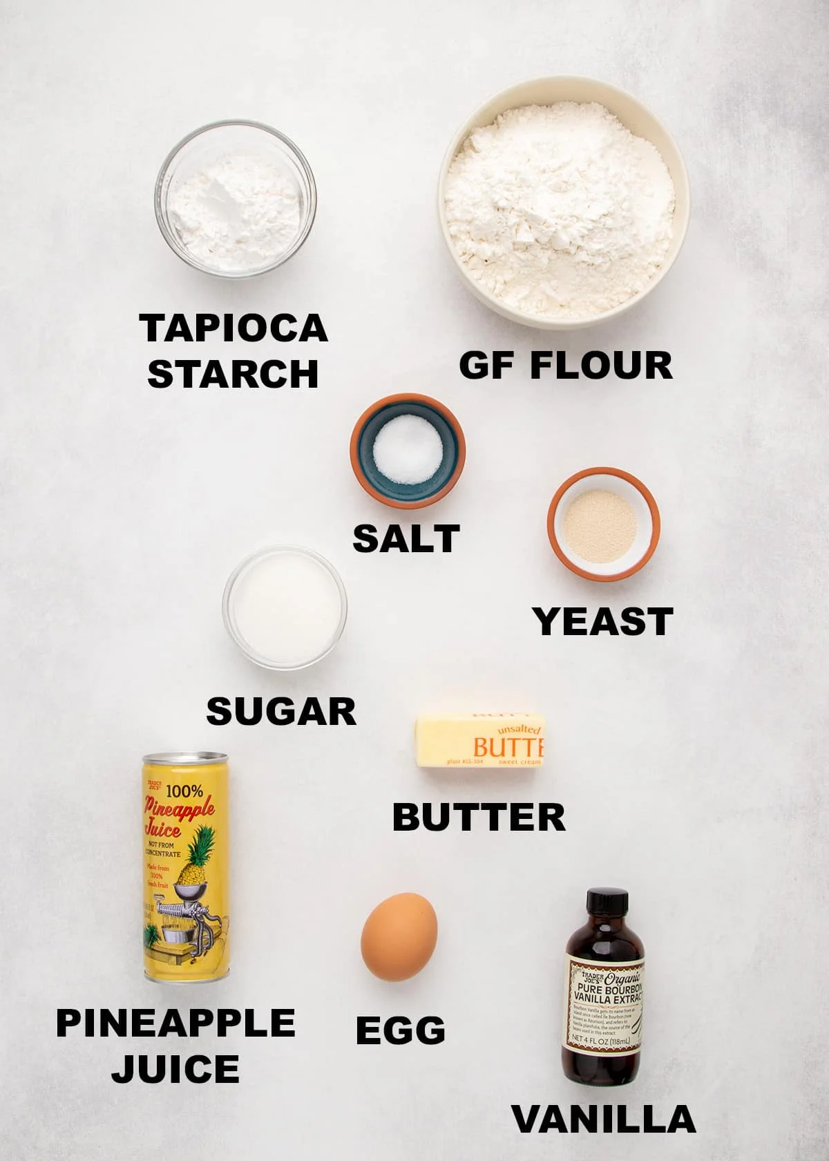 Ingredients for gluten free Hawaiian rolls arranged on a light surface labeled tapioca starch, gluten free flour, salt, yeast, sugar, butter, pineapple juice, egg, and vanilla.