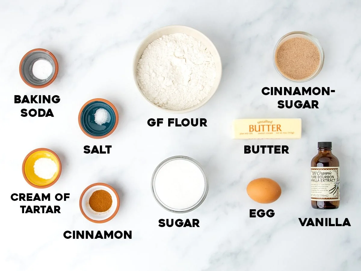 ingredients like gluten free flour, cream of tart, sugar, cinnamon, egg, vanilla and butter for gluten free snickerdoodles cookies on marble surface with names of ingredients