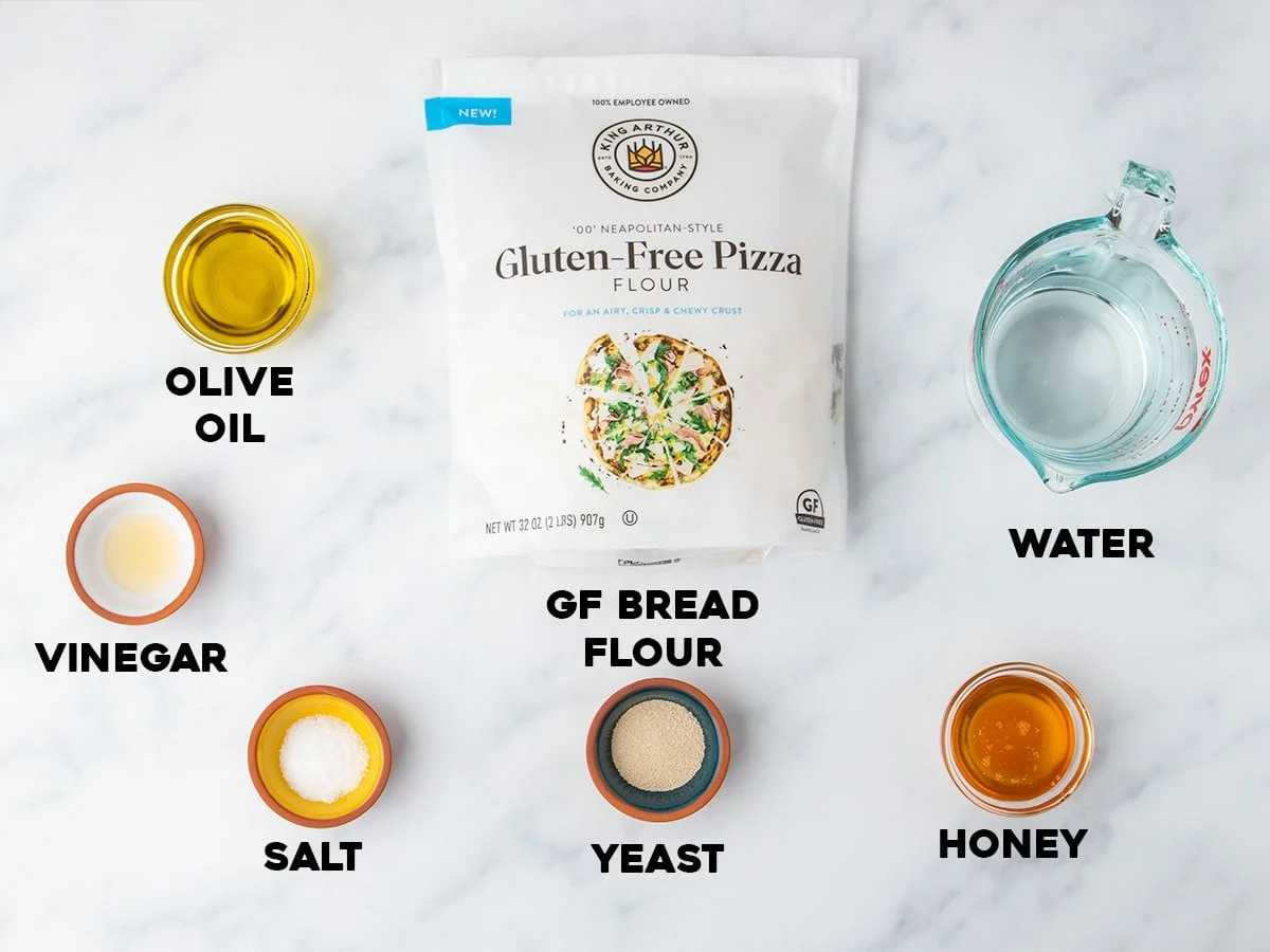 Labeled ingredients displayed on a white surface, including bag of gluten free flour, olive oil, honey, yeast, water, vinegar, and salt.