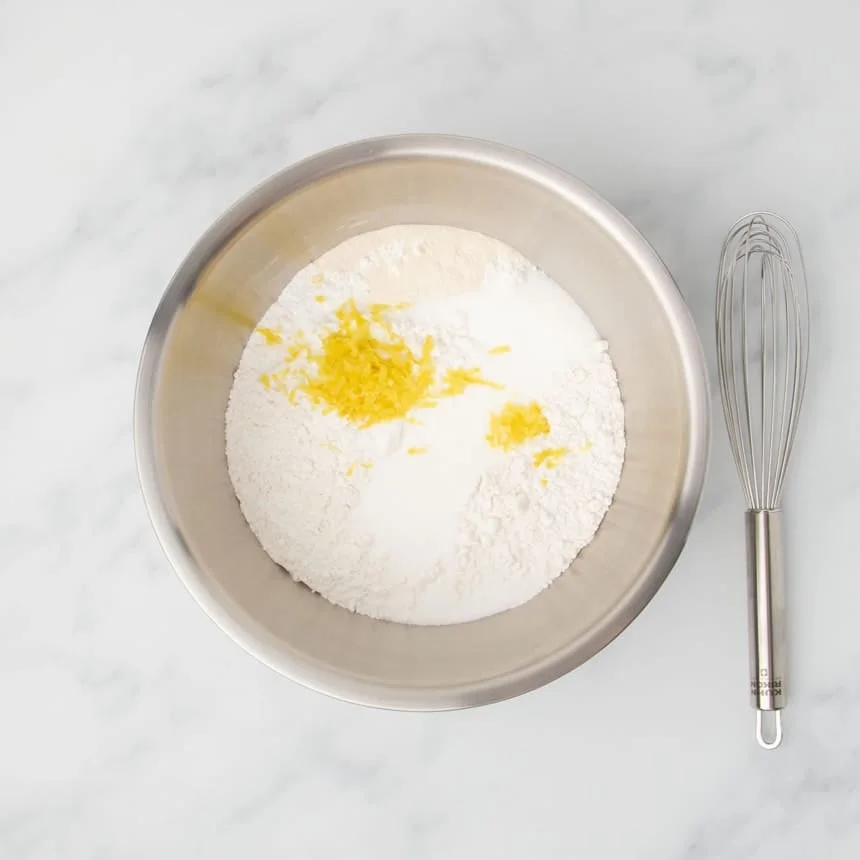 Large bowl with dry ingredients including flour, sugar, and yellow shredded lemon zest, with a whisk resting next to the bowl.