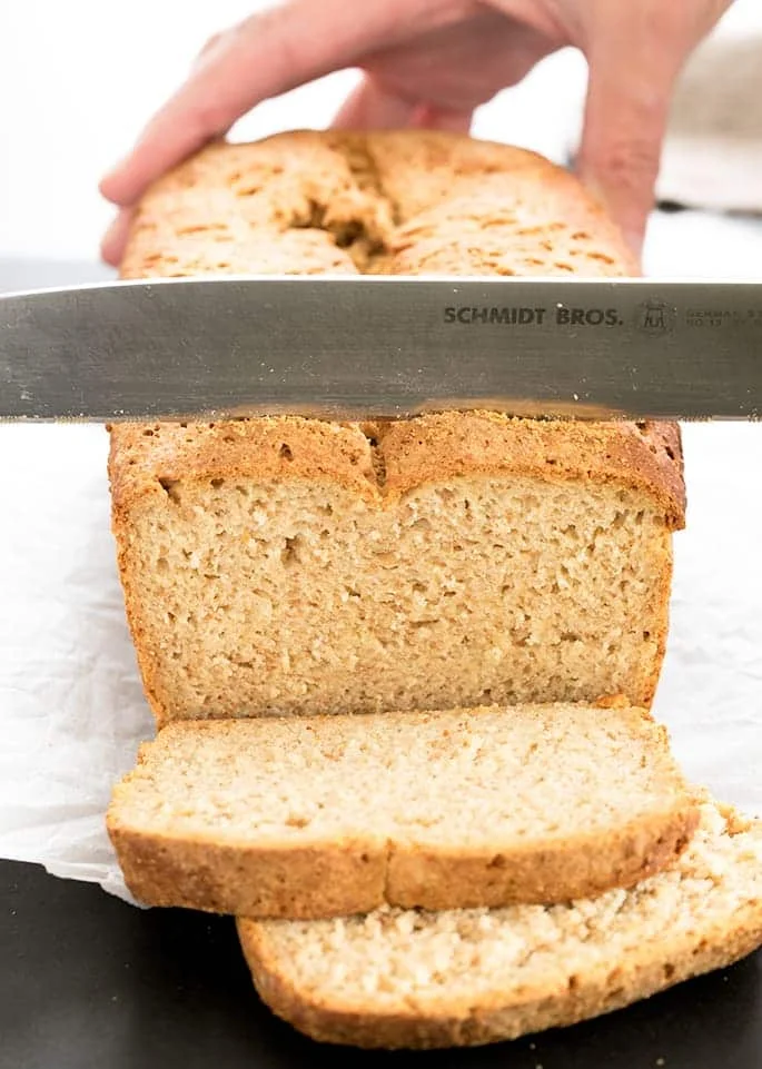 large bread knife cuts brown bread loaf into slices while other hand holds the bread in place on white paper with crumbs