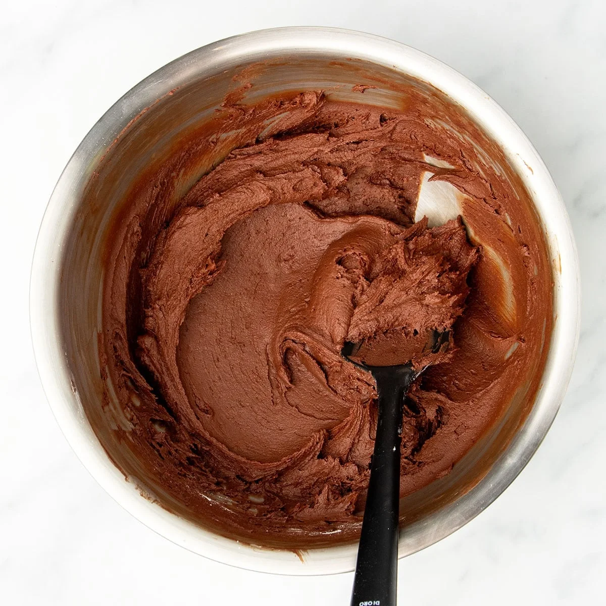 large round metal mixing bowl with dark brown chocolate frosting with black silicone spatula