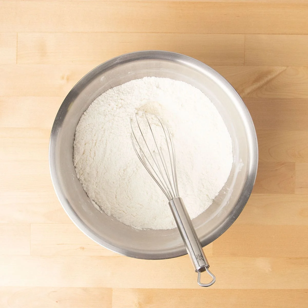Large round metal mixing bowl with dry ingredient and wire whisk.