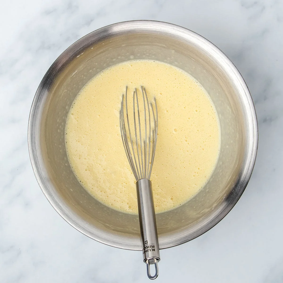 large round metal mixing bowl with frothy light yellow liquid mixture and metal wire whisk