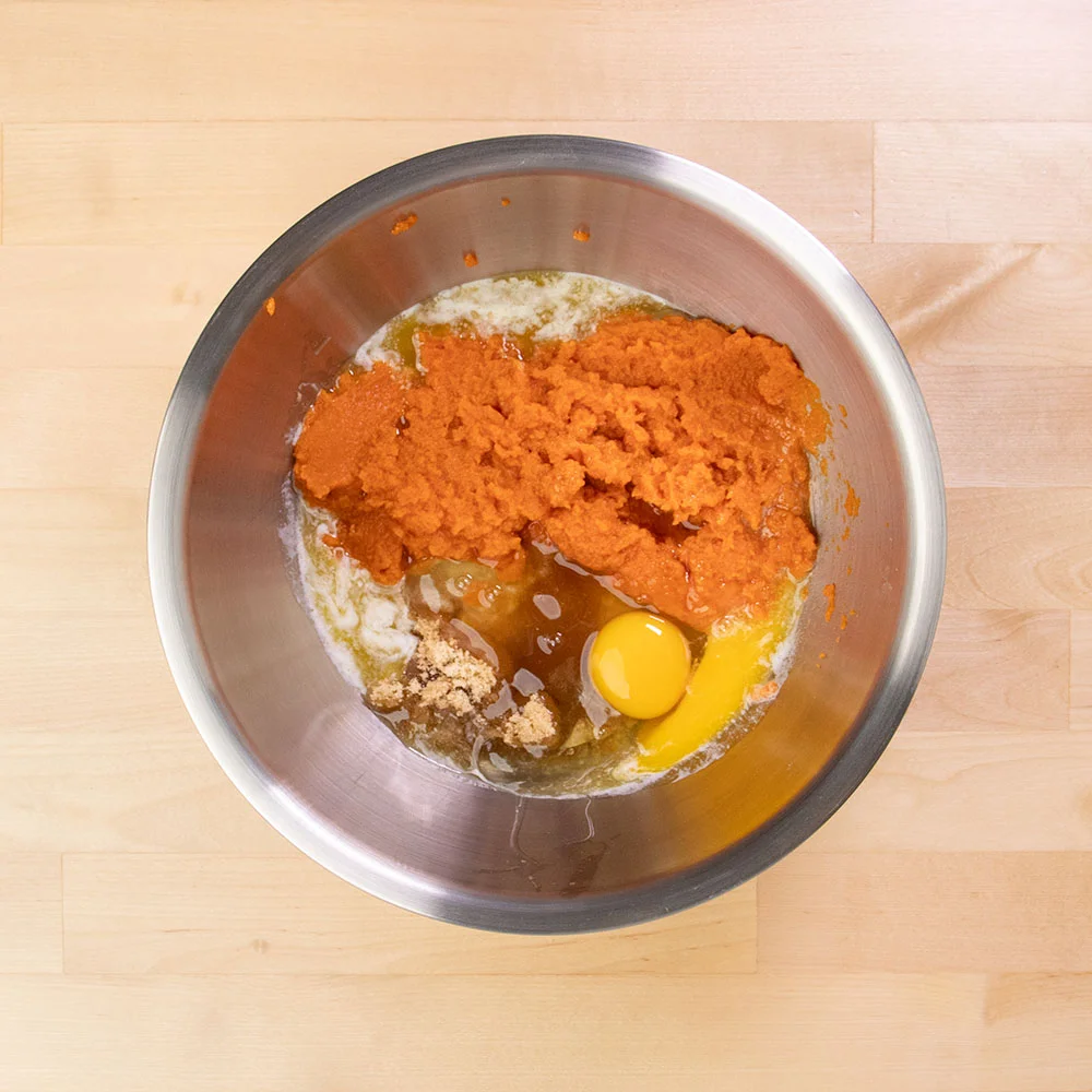 large round metal mixing bowl with orange pumpkin, yellow eggs, melted butter and sugar