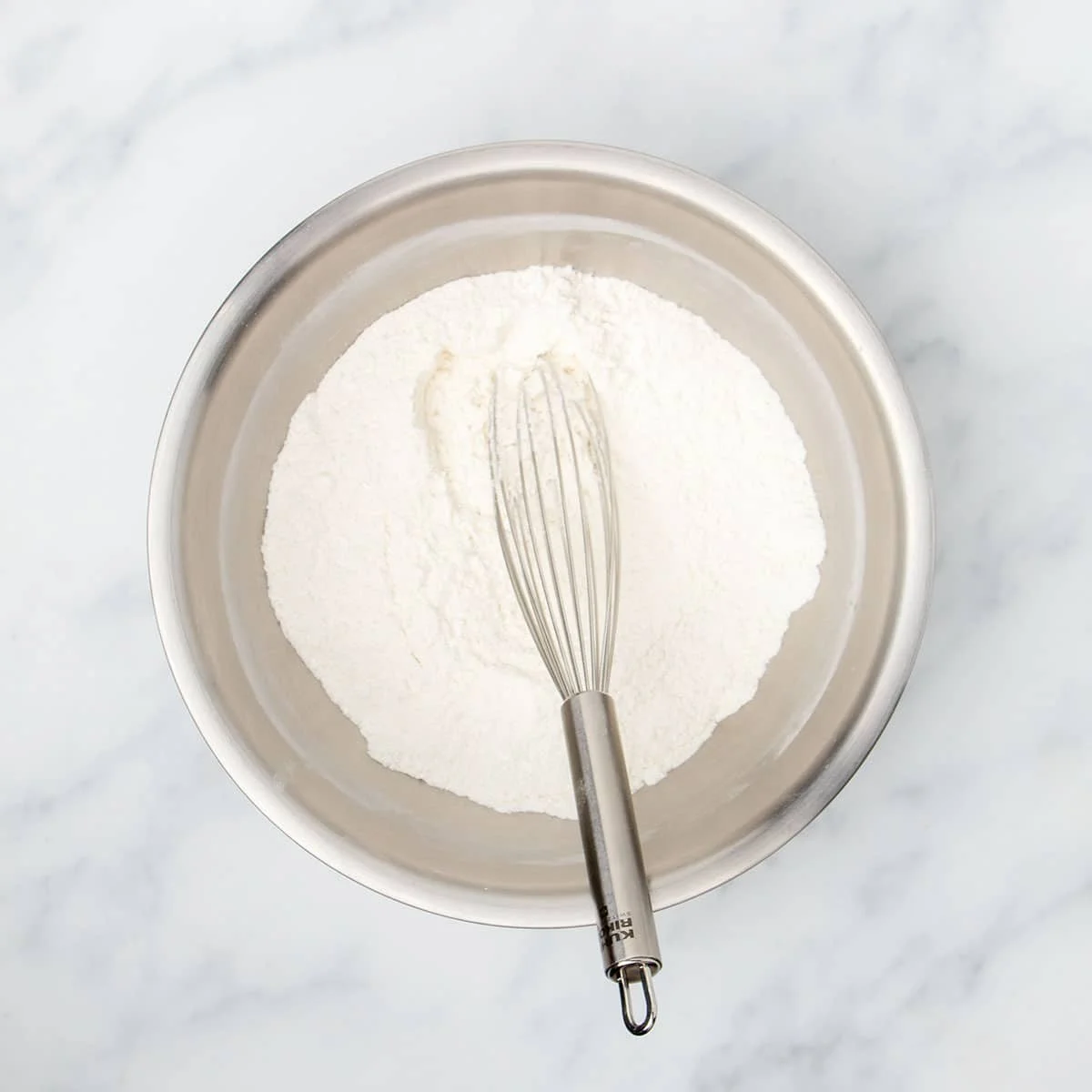 Large round metal mixing bowl with white flour mixture and large metal wire whisk.