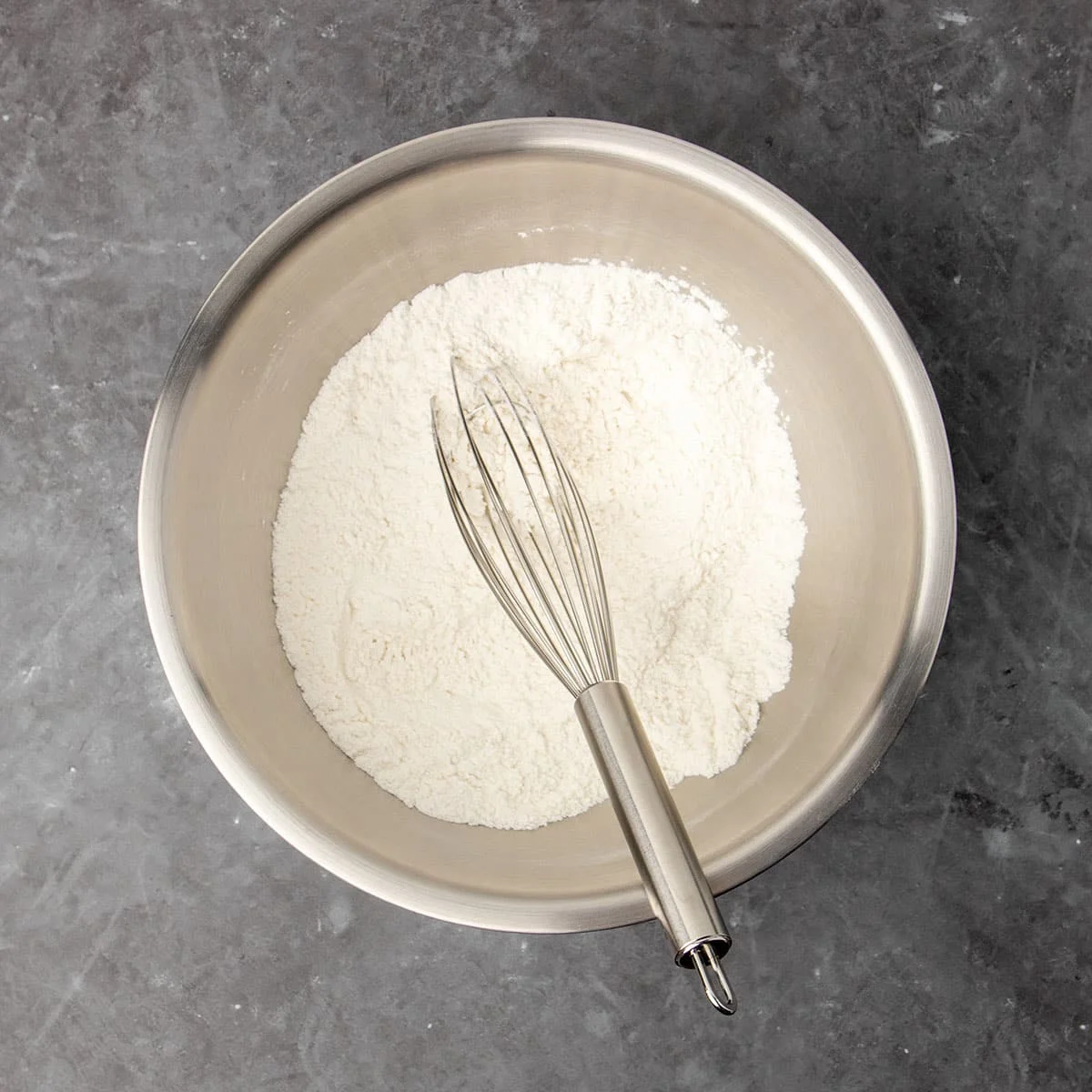 Large round metal mixing bowl with white flour mixture and metal wire whisk