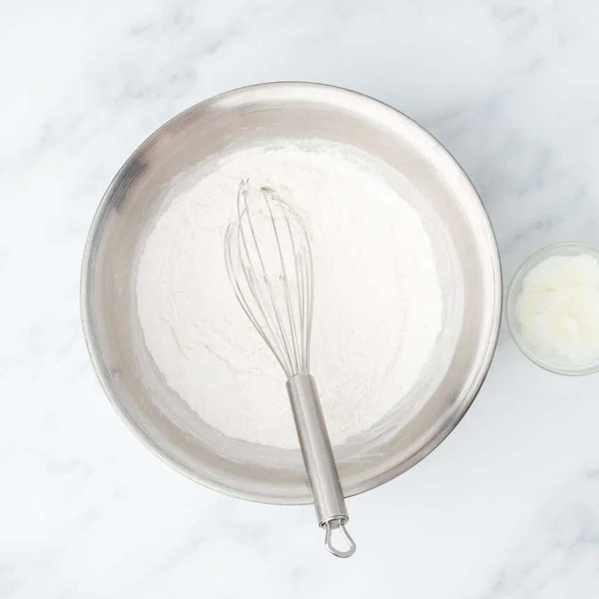 large round silver colored mixing bowl with white flour mixture and large metal wire whisk