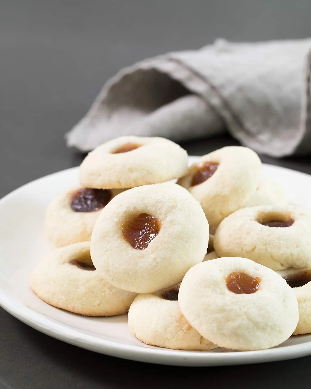 large round white serving platter with a pile of small round thumbprint butter cookies with dark or apricot colored jam in the center