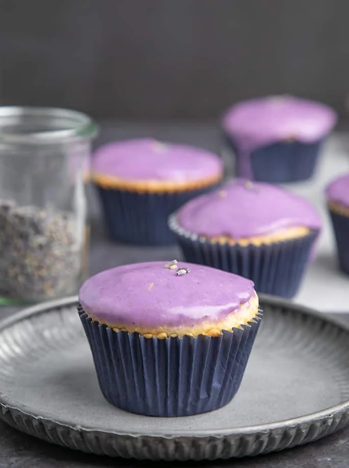 Lavender glazed cupcake on small metal plate with more in background.